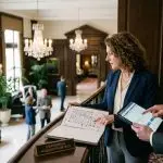 Two business professionals review a binder with a floor plan and a tablet in a luxurious lobby, a sign reads 'Confidential Acquisition Details'.